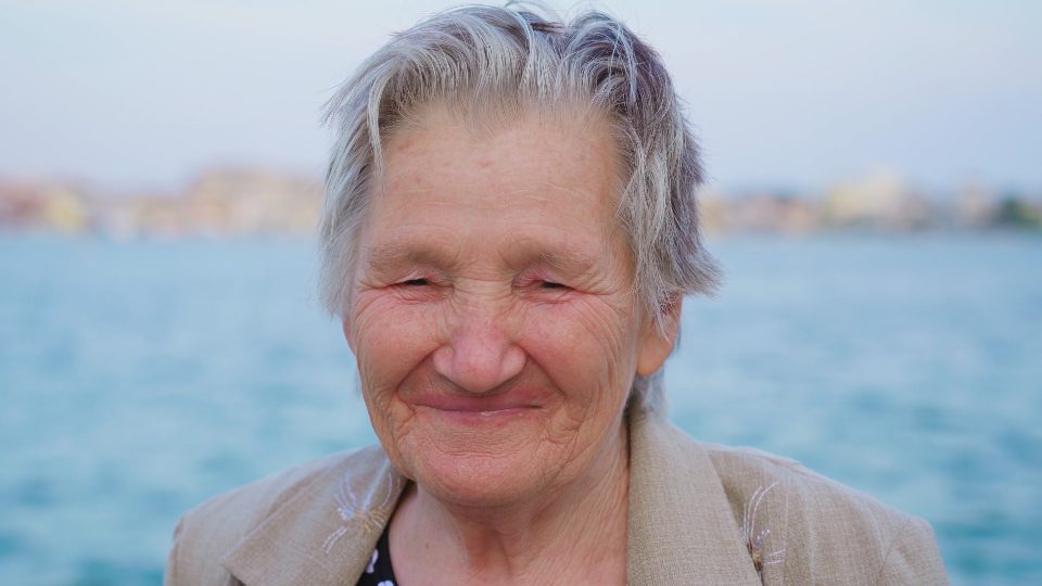Senior grey haired woman traveler with wrinkled face laughs happily standing on promenade in summer evening in Chioggia extreme closeup