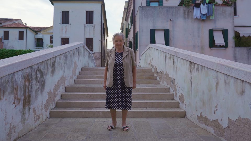 Senior woman traveler with grey hair stands on city stone footbridge smiling among historical buildings in Chioggia in summer after sunset