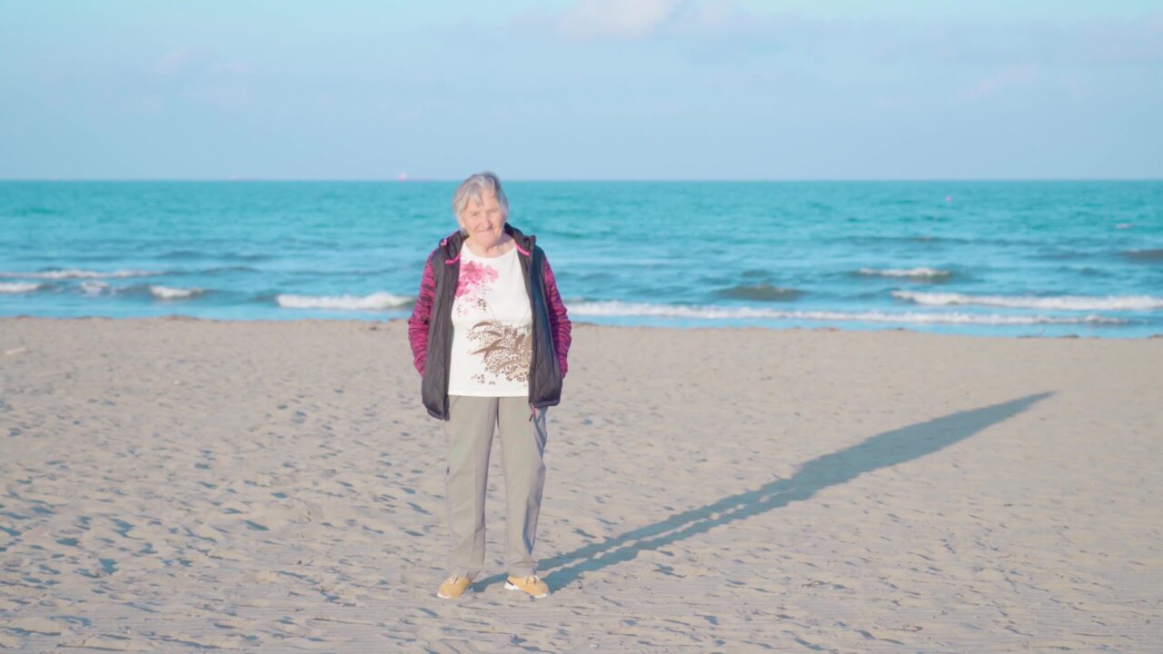 Elderly woman stands on sea beach against waves at sunset