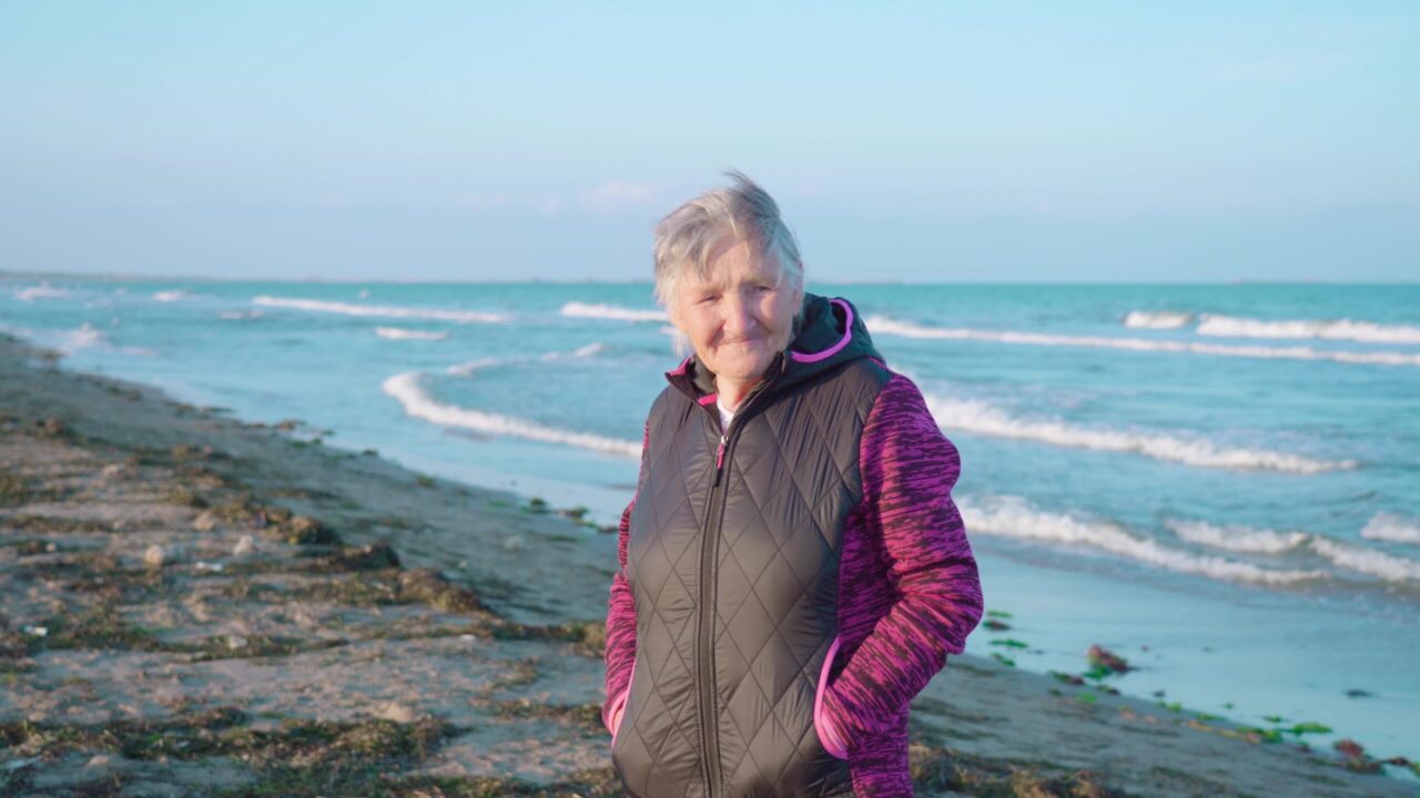 Senior grey haired woman with short haircut in windbreaker enjoys weather standing on coastal beach edge against sea closeup