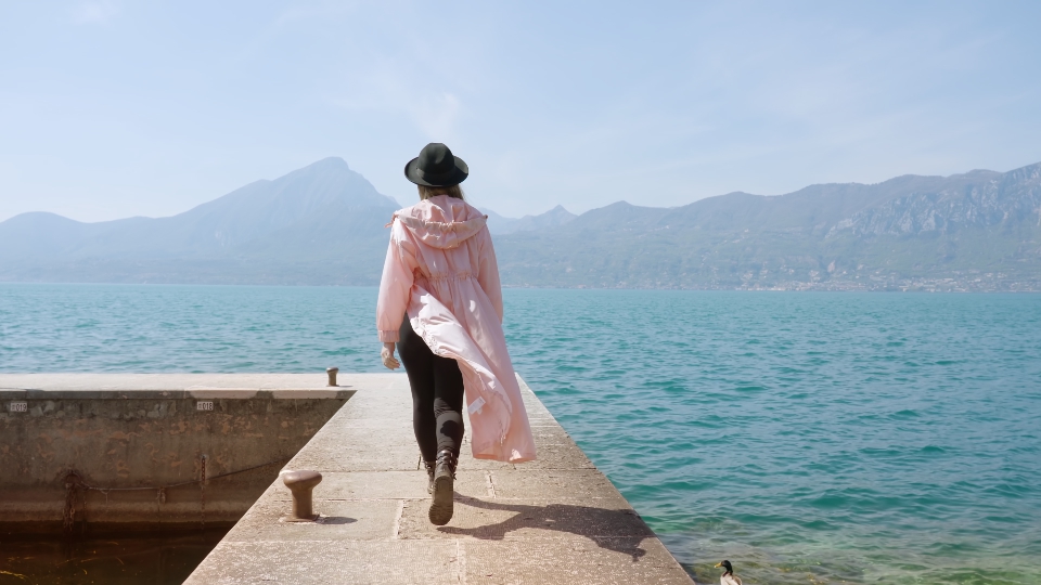 Woman in coat walks on pier in Lake Garda with blue water