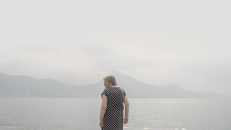 Elderly woman in polka-dot dress stands on lake waterfront