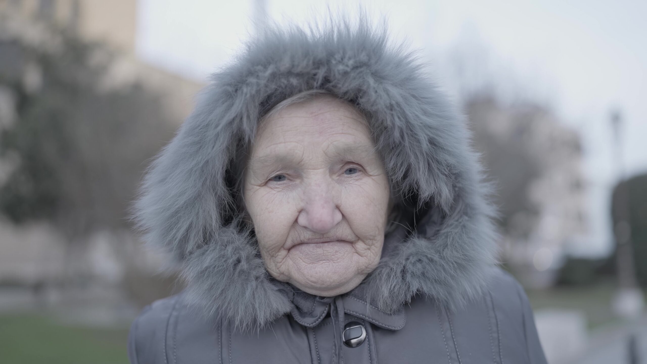 Elderly woman in fur hooded jacket stands outside building