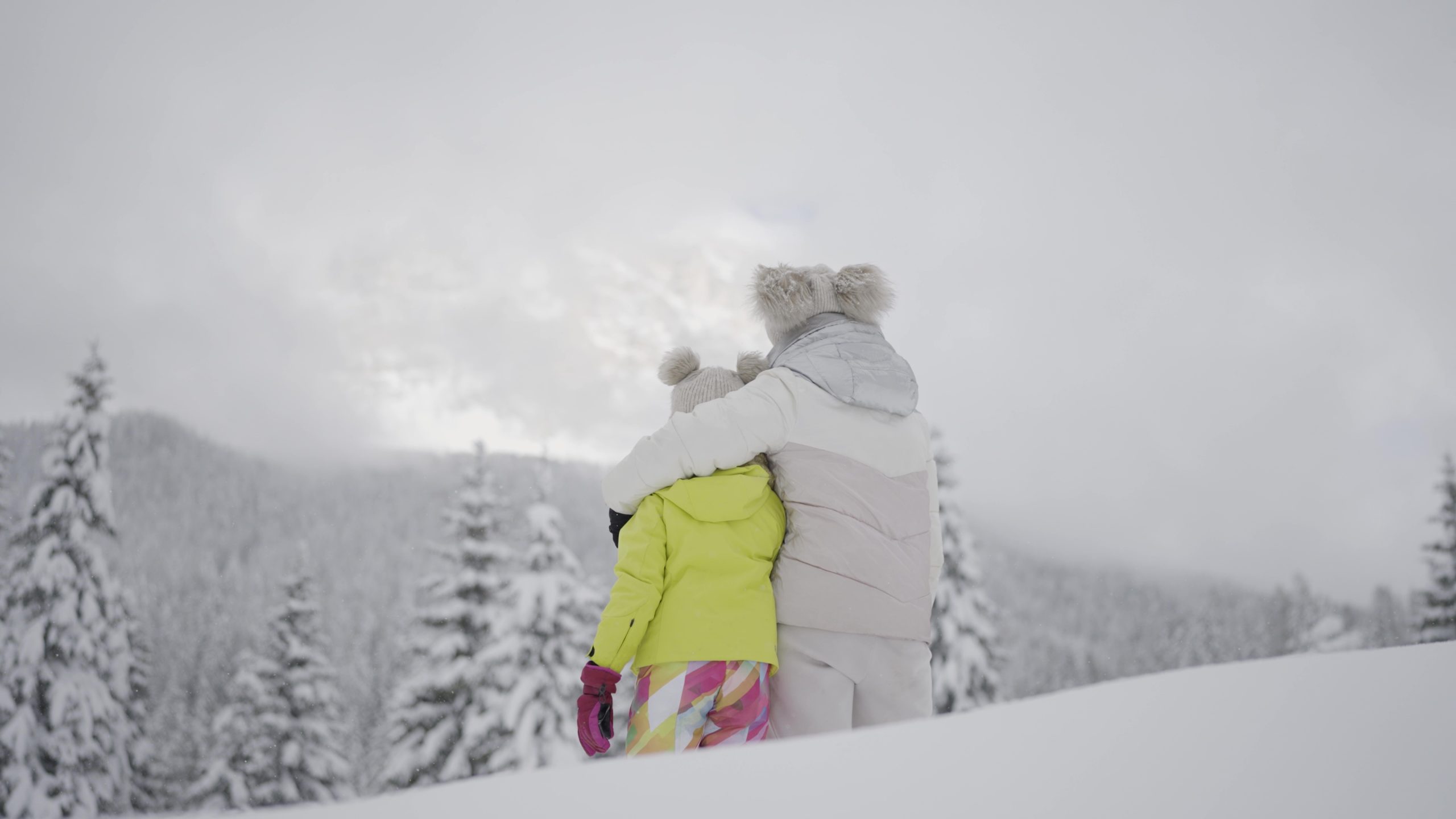 Mom and daughter look at forest in the snow under high mountains